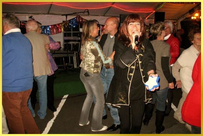 Schlagerpiratin singt in die Kamera und lacht. Partyg�ste dahhinter tanzen.